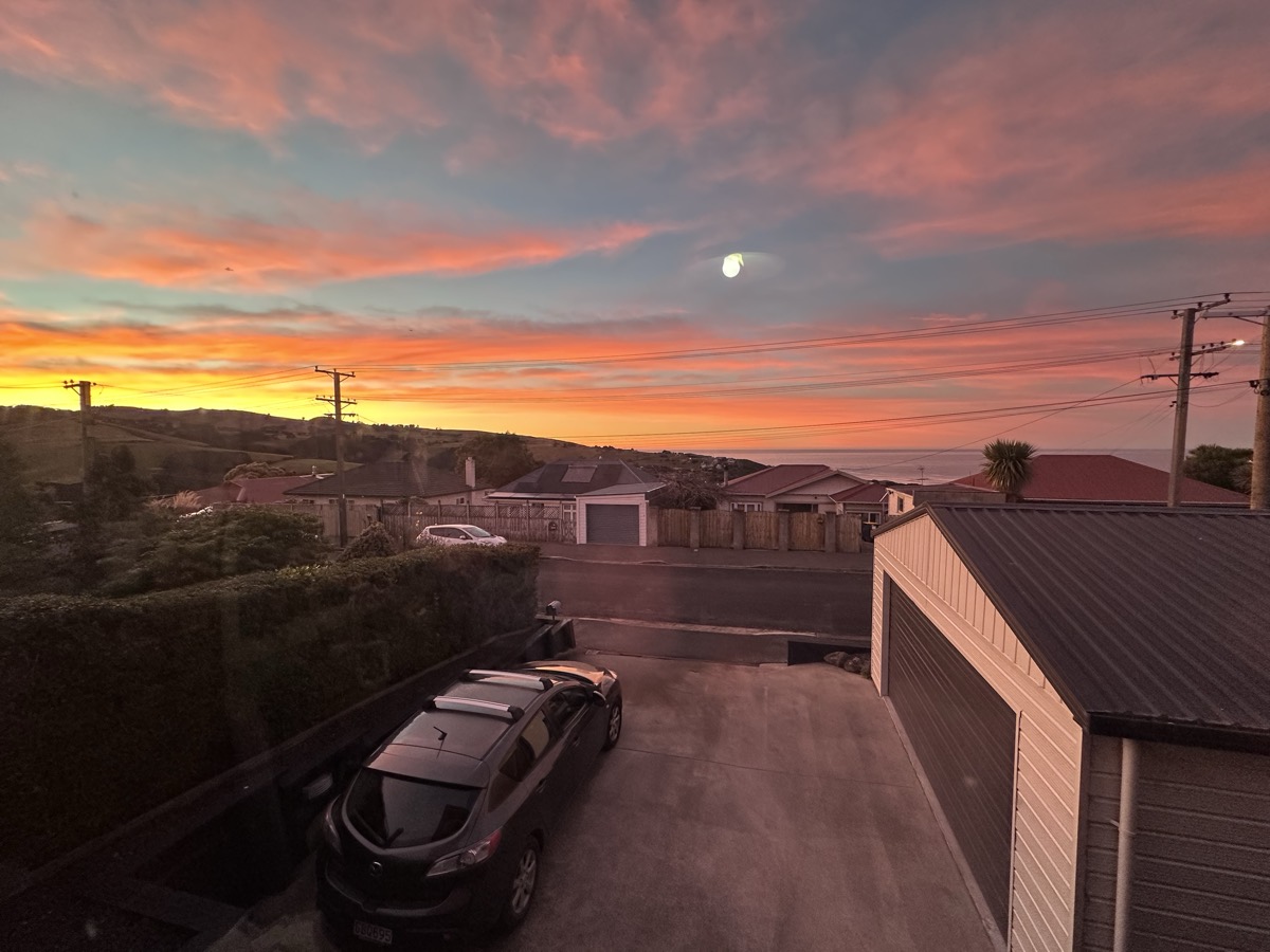 Evening view over the rooftops of a Dunedin suburb toward the hills and sea, with the moon rising